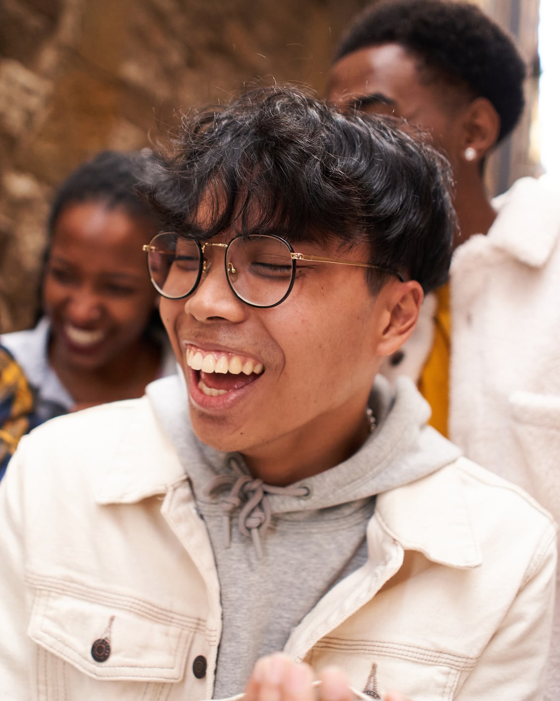 A young person wearing glasses is smiling with two other young people in the background