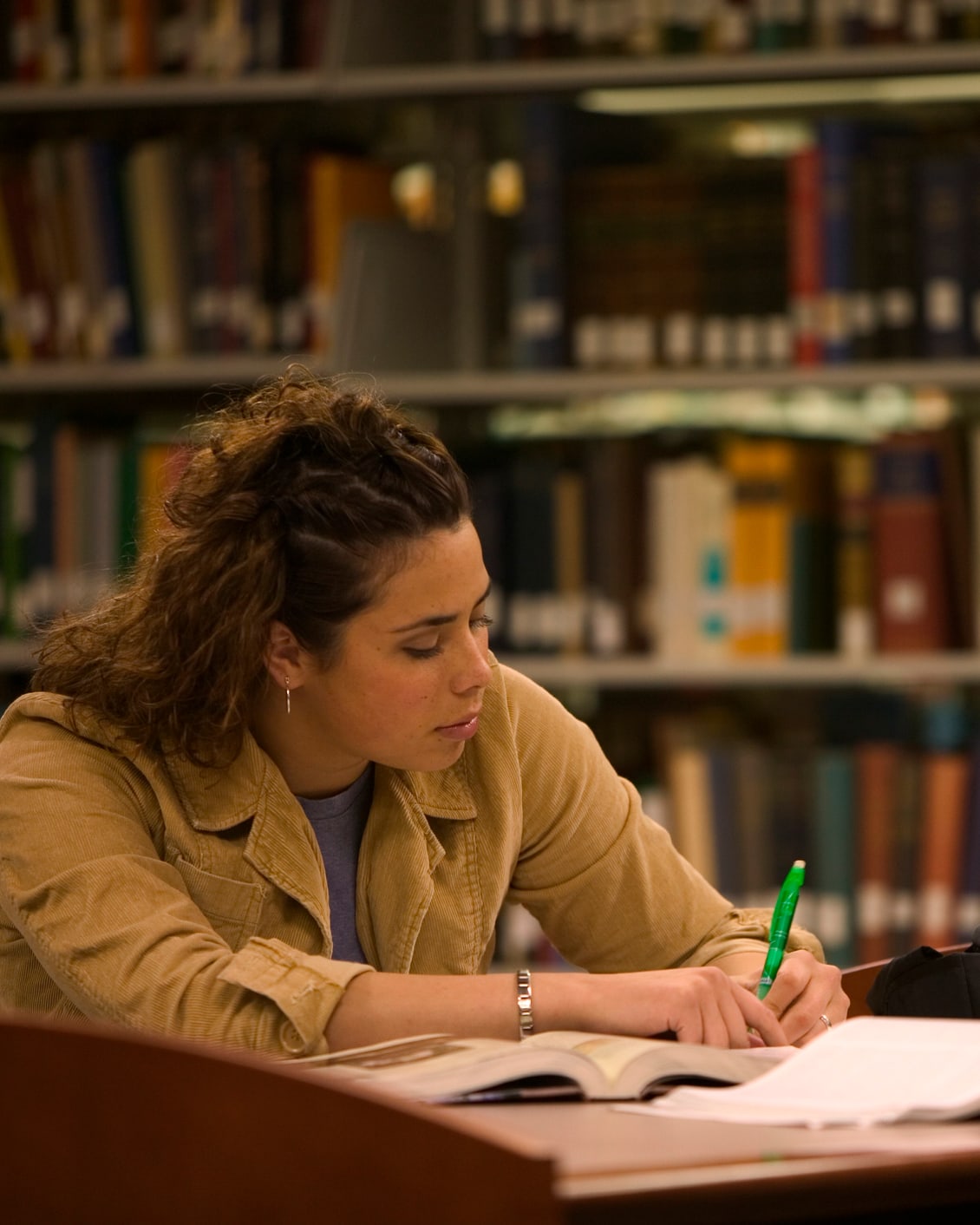 Young woman in library is writing in a book with a green pen.